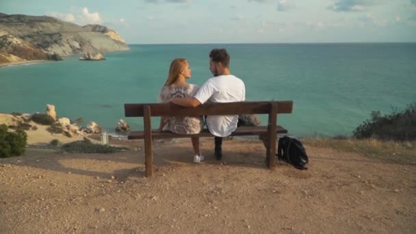 Jovem casal abraço está sentado em um banco em um penhasco com uma vista pitoresca lindo do mar sem fim com água limpa — Vídeo de Stock