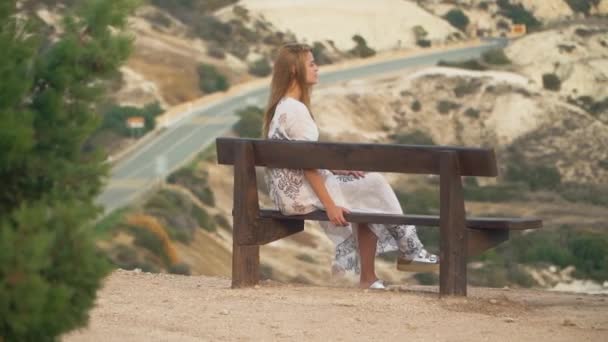 Μοναχικό κορίτσι σε μακρύ λευκό φόρεμα που κάθεται στον πάγκο. Όμορφες βραχώδεις λόφους και δρόμο που εκτελούνται στο παρασκήνιο. Laisure ξανθιά γυναίκα στο καλοκαίρι. Κάμερα κινείται προς τα αριστερά — Αρχείο Βίντεο