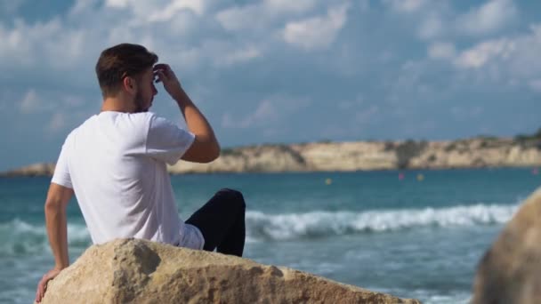 Jovem homem bonito sentar-se na pedra perto do mar incrível — Vídeo de Stock