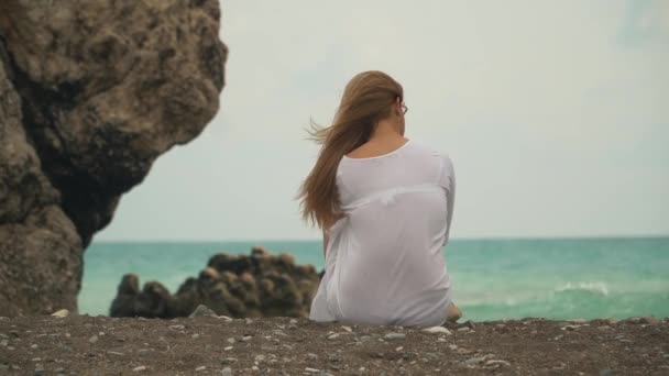 Красивая девушка сзади с красивыми светлыми волосами бросает камешки в воду на фоне живописного синего волнистого моря — стоковое видео