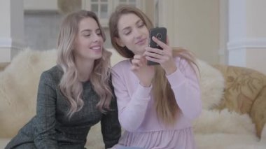 İki genç ve güzel kız moda elbiseler seyir telefon oturma odasında kanepede oturuyor. Kızlar iyi indirimler arayışı içinde.