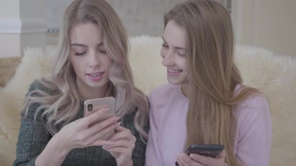 Twee mooie jonge vriendinnen zittend op de Bank in de woonkamer op zoek naar goede kortingen voor het winkelen via internet in hun gadgets. Winkelen op concept. — Stockvideo
