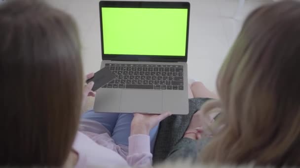 Две девушки ищут хорошие скидки на покупки с помощью Интернета на их ноутбуке с зеленым экраном сидит на диване в гостиной. Одна девушка держит кредитную карту в руке, готовую к — стоковое видео