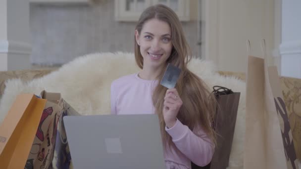 ブラック クレジット カードを表示するノート パソコンと近く、ショッピング バッグの多くが付いているソファーに座っているかわいい女性。女の子はオンラインで多くのものを買った。美しい女性の買い物に成功 — ストック動画