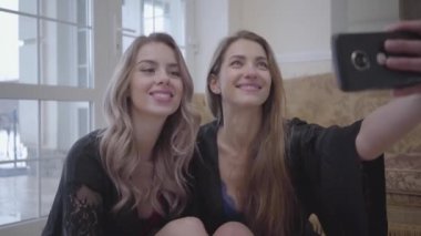İki güzel kadın evde katta oturan ve selfie cep telefonu alarak siyah elbiseli kapatın. Kızlar iyi bir fotoğraf için poz. Modern kadınların eğlence