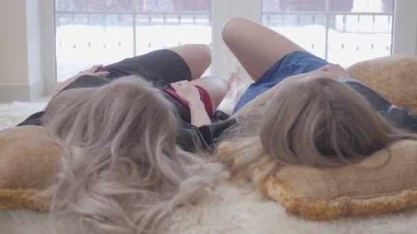 Two beautiful girls lying in bed close up, looking up and kicking each other with legs. Girlfriends dressed in nighties and black robes rest at home. Concept of women friendship. — Stock Video