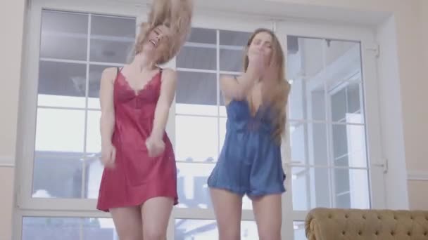 Dos hermosas chicas jóvenes bailando en la sala de estar moderna cerca de grandes ventanales por la mañana. Las novias vestidas con camisas rojas y azules se divierten en casa. Concepto de mujer — Vídeo de stock