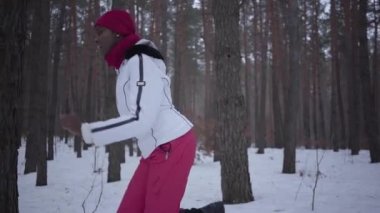Afrikalı-Amerikalı kadın kırmızı şapka, eşarp ve karlı ormanda çalışan beyaz ceket giyiyor sıcak giyinmiş. Kız sürekli geri çalışan takipçisi uzak görünüyor. Ağır çekim.
