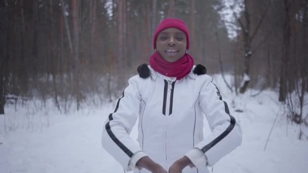 African american girl werpt een handvol sneeuw staan in winter bos en kijken in de camera. Beartiful meisje in warme jas tijd buitenshuis doorbrengen. Concept van openluchtrecreatie — Stockvideo