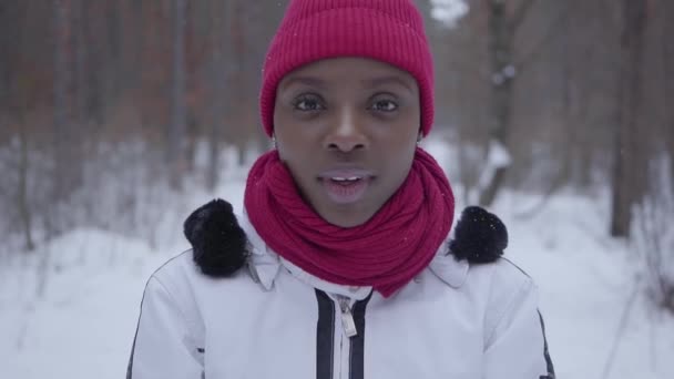 アフリカ系アメリカ人の少女の肖像画を一握り雪冬の森に立って、カメラで見ているをスローします。屋外で過ごす時間の暖かいジャケットの美しい少女。屋外レクリエーションの概念 — ストック動画