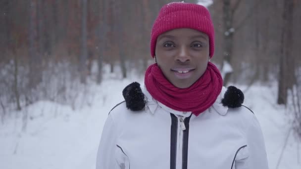 Portrait confident successful young smiling African American girl dressed warm wearing a red hat, scarf and white jacket standing in a snowy forest outside the city. Real people series. Slow motion. — Stock Video