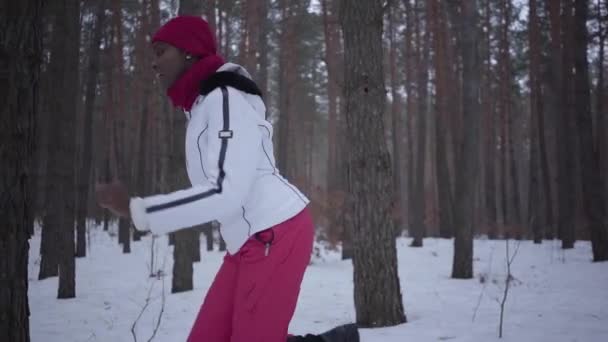 Afroamerikanska kvinna klädd varma bär en röd hatt, halsduk och vit jacka kör genom den snöiga skogen. Flickan ser ständigt tillbaka körs bort från förföljaren. Slow motion. — Stockvideo