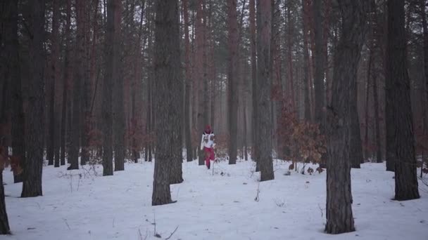 Chica afroamericana corriendo a través del bosque de invierno hacia la cámara, sonriendo. Hermosa chica en chaqueta caliente se detiene a descansar bajo el árbol. Concepto de actividad al aire libre. Disparo en cámara lenta — Vídeos de Stock