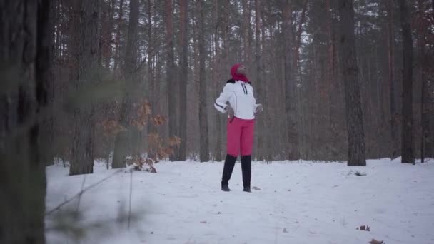 Fata afro-americană pierdută îmbrăcată caldă purtând o pălărie roșie, eșarfă și jachetă albă în picioare în pădurea de iarnă și se uită speriată în jur . — Videoclip de stoc