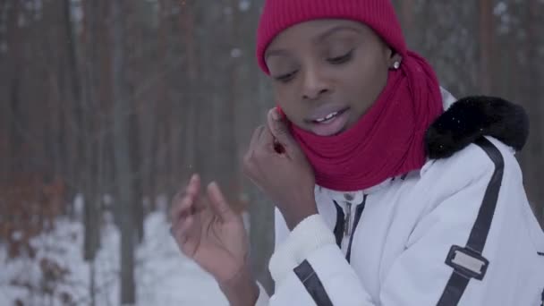 Afrikansk amerikansk pige taler via mobiltelefon stående i vinterskoven tæt op. Smuk pige i hvid jakke vender sig og ser bag, hun er bange. Kvinde forsøger at kalde hjælp – Stock-video