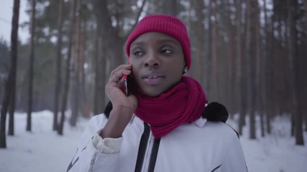 Retrato de menina afro-americana falando por telefone celular na floresta de inverno sorrindo de perto. Menina bonita em casaco quente passar tempo ao ar livre. Câmera se move para a esquerda — Vídeo de Stock