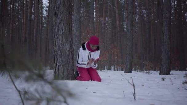 Chica afroamericana asustada llamando por teléfono celular sentada detrás de un árbol en el bosque de invierno. Mujer tratando de llamar a ayuda — Vídeos de Stock