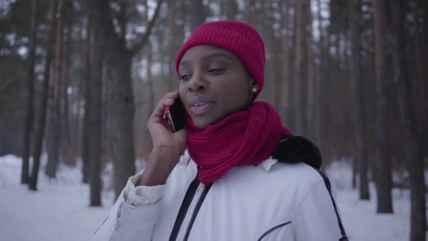 Retrato joven afroamericano chica hablando por teléfono celular en el bosque de invierno sonriendo de cerca. Hermosa mujer bonita vestida caliente con un sombrero rojo, bufanda y chaqueta blanca pasar tiempo al aire libre . — Vídeos de Stock