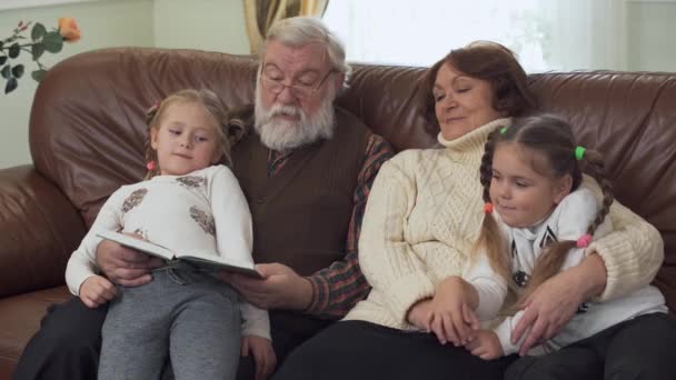 Abuelo y abuela sentados en un sofá de cuero en la moderna sala de estar con dos niñas pequeñas cerca. Hombre adulto leyendo libro para sus nietas. Ocio de familia feliz — Vídeo de stock