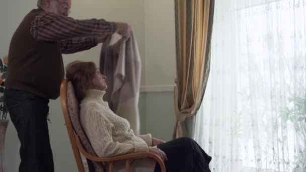 成熟的女人坐在现代房子大窗户附近的摇椅上。她的丈夫来给妻子盖上毯子, 然后亲吻她的脸颊。老年夫妇彼此相爱和支持 — 图库视频影像