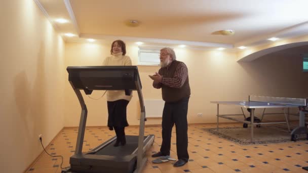 Senior woman running on treadmill on toes in big light room at home. Mature bearded man standing near and applouds her. Senior couple love and support each other — Stock Video