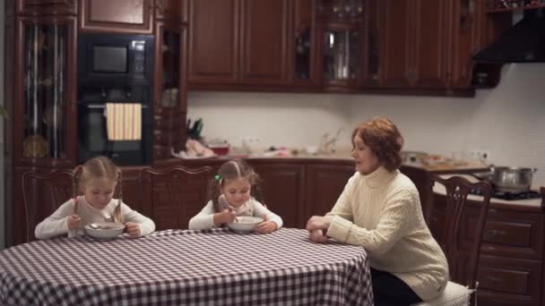 Abuela y dos preciosas nietas sentadas en un gran kitcken en la mesa comiendo sopa. Familia feliz almorzar en casa. Mujer mayor alimentando a niños pequeños — Vídeos de Stock