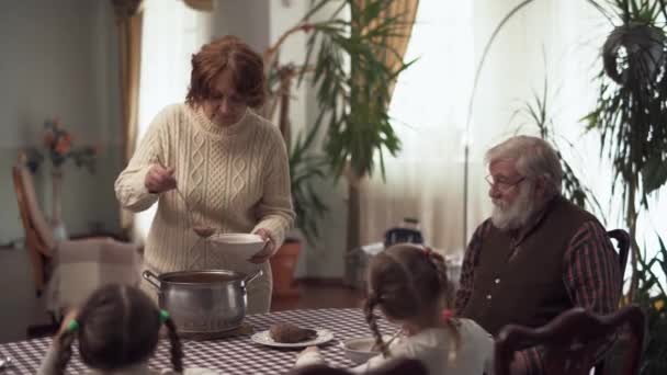 Бабушка наливает борщ в миски для мужа и двух милых внучек, пока они сидят и ждут — стоковое видео