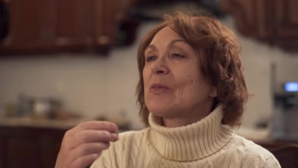 Starší žena v teplém svetru jí sladkosti sedí doma v moderní kuchyni, pak ukazuje velký prst. Zralá žena se těší její oběd — Stock video