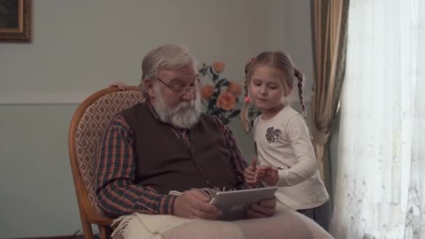Γενειοφόρος ανώτερος άνθρωπος κάθεται σε κουνιστή καρέκλα με tablet στα χέρια. Δύο μικρά κορίτσια στέκεται κοντά παππού και ψάχνει το gadget του. Τα παιδιά έχουν μια διασκέδαση στο σπίτι οι παππούδες — Αρχείο Βίντεο