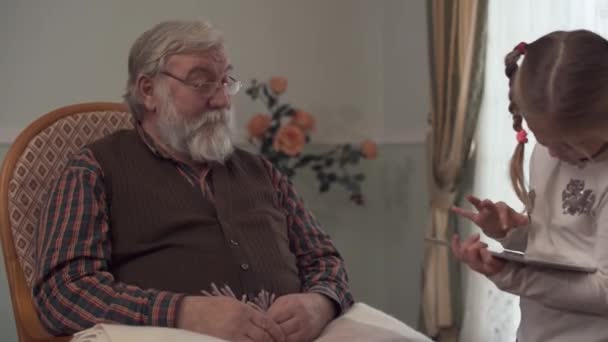 Bearded senior man sitting in rocking chair near big window looking at his granddaughter. Small girl standing near grandfather and playing on tablet. Kid having fun in grandfathers house — Stock Video