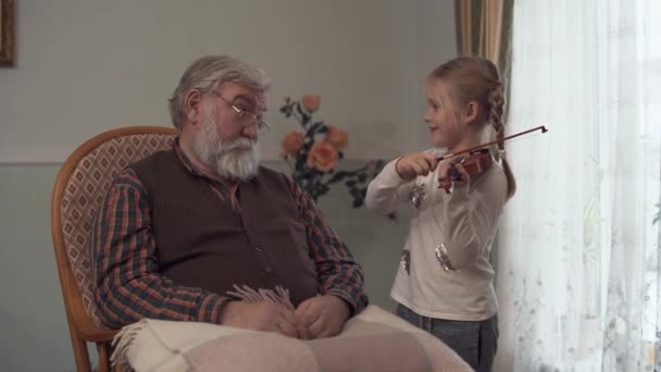 Grandfather sitting with plaid on the rocking chair whil his funny granddaughter dabbling and plays the violin badly — Stock Video
