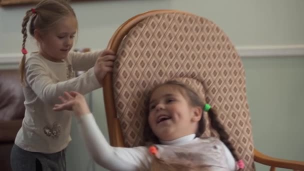 Petite fille secouant sa sœur dans un fauteuil à bascule de près. Deux jolies filles avec des tresses s'amusent ensemble à la maison. Les sœurs sont amicales — Video