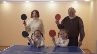 Oyun salonu yakınındaki Tenis tablo iki küçük granddaughters, tüm sallayarak raketleri, sonra dalgalar eller ile ayakta dedesi. Bir kız surat. Mutlu aile eğlence
