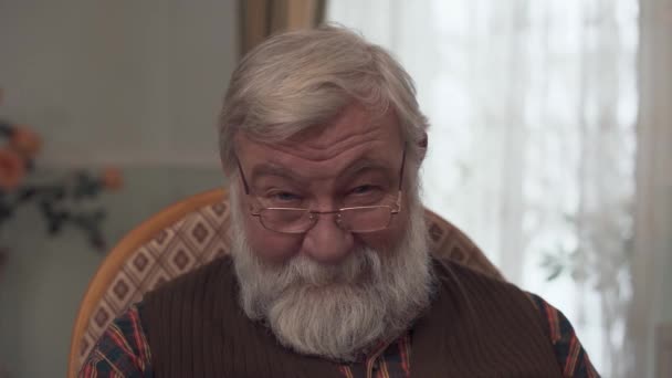 Portrait of an adult smiling grandfather with a big gray beard and a good look who wears glasses sitting on a rocking chair and looking at the camera. Adult man wearing a red vest and a plaid shirt — Stock Video