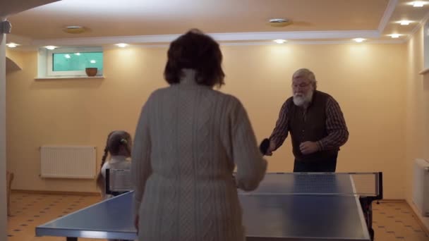 Dědeček a babička si zahrát stolní tenis, dvě malé dívky sledují hru. Starší žena dává průchod k muži, ho nemotorně honí míč. Volný čas šťastná rodina — Stock video