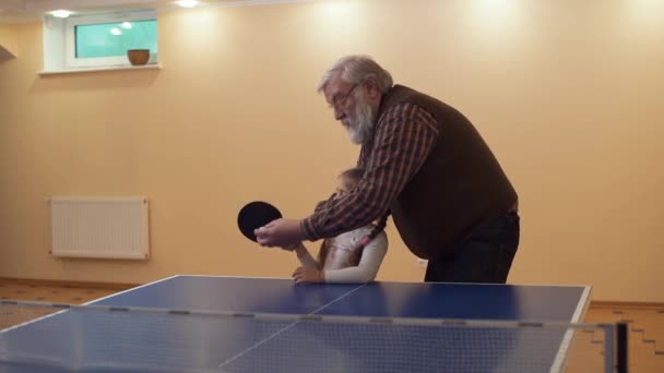 Grandfather teaches his little cute granddaughter to play tennis and helps her make a punch. Slow motion — Stock Video