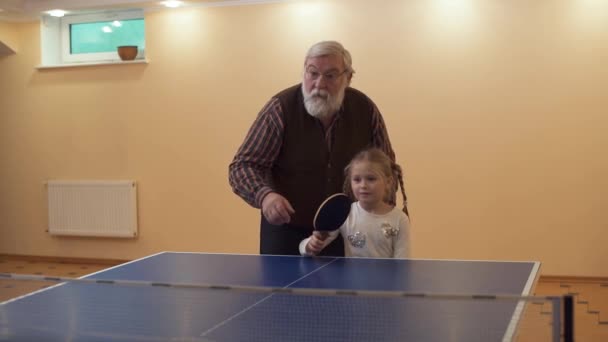 El abuelo enseña a su nieta a jugar al tenis de mesa, de pie junto a ella. La niña golpea la pelota. Ocio de familia feliz — Vídeos de Stock