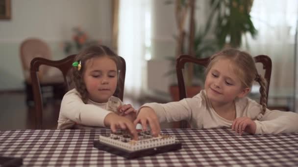 Dos chicas lindas con coletas toman dulces de la caja de caramelos y se muestran el uno al otro sentados en la mesa de la cocina. Hermanas comiendo chocolate en casa — Vídeos de Stock