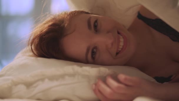 Милая молодая женщина с рыжими длинными волосами лежит в постели и улыбается. — стоковое видео