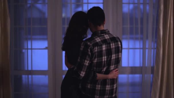 Man and woman standing near floor-to-ceiling window looking outside at home. Happy couple cuddle each other. Shooting from behind — Stock Video