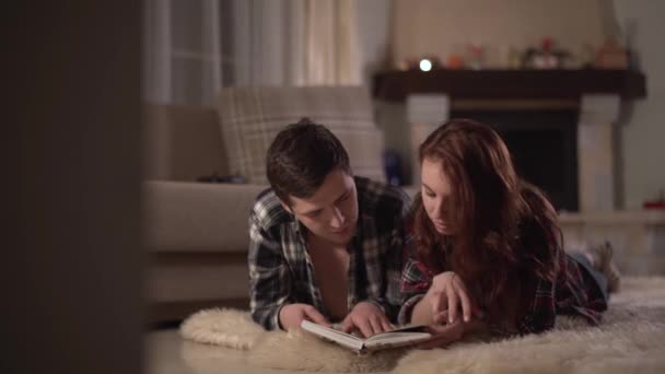 Fiatal, boldog férfi és nő feküdjön le bolyhos curpet, a nappaliban kandalló és egy könyvet olvas. Egy fiatal pár a szerelem időtöltés. Szabadidő, a fiatal család. — Stock videók