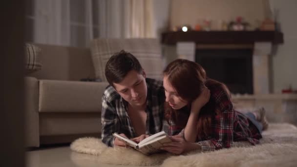 Young happy man and woman lie down on fluffy curpet in the living room with fire place and reading a book. Pastime of a young couple in love. Leisure of young family. — Stock Video
