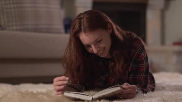 Mulher bonito em camisa xadrez deitado no tapete fofo lendo o livro de perto. Lady vira páginas e sorri, relaxando em casa. Lazer de mulher jovem à noite — Vídeo de Stock