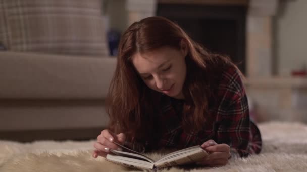 Mulher bonita em camisa quadriculada deitada no tapete fofo lendo o livro de perto. Lady vira páginas e sorri, relaxando em casa. Lazer de mulher jovem à noite — Vídeo de Stock