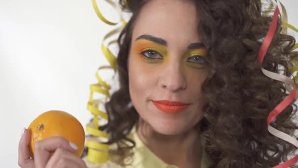 Gros plan portrait de jeune fille souriante ludique avec un maquillage lumineux tenant orange. Mouvement lent . — Video