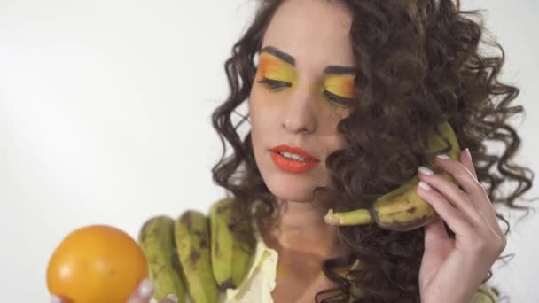 Портрет молодої кучерявої дівчини з яскравим макіяжем, що дивиться на помаранчевий з інтересом імітує телефонну розмову з бананом. Повільний рух — стокове відео