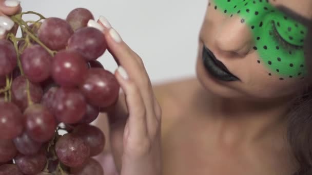 Close up portrait of cute young curly girl with creative make up tenderly holding a bunch of ripe grapes — Stock Video