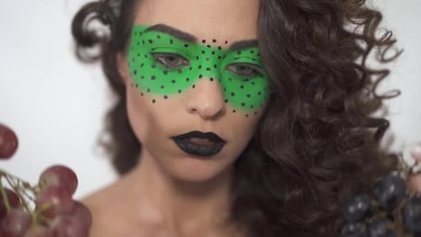 Portrait of beautiful curly girl with creative make up seriously choosing between two ripe bunches of grapes — Stock Video