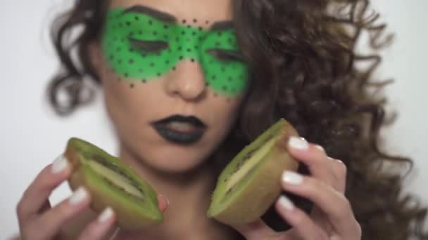Portrait beautiful smilling curly girl with bright make up holding two halves of kiwi. — Stock Video