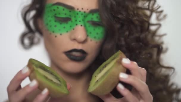 Porträt schöne lächelnde lockige Mädchen mit hellem Make-up hält zwei Hälften Kiwi. — Stockvideo